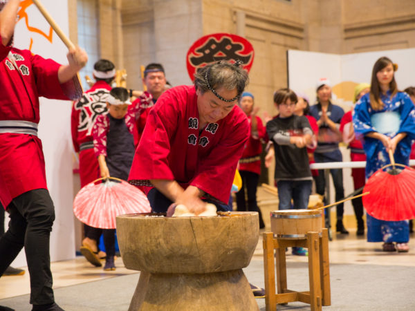 A man wearing a red tunic and black headband kneads mochi in a tall wooden bowl.