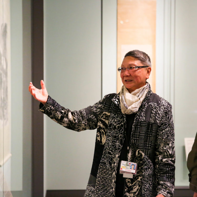 A docent gestures towards an artwork while a visitor looks on.