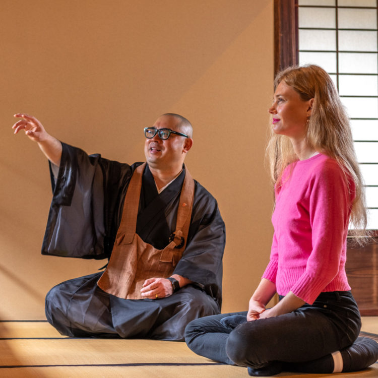 Seated man wearing robes points into middle distance while a kneeling woman in a pink sweater looks on.
