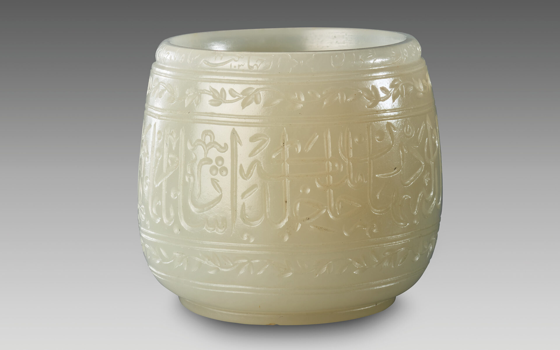 A small, shiny, cream-colored jade cup with Arabic script and flowers carved on the surface.