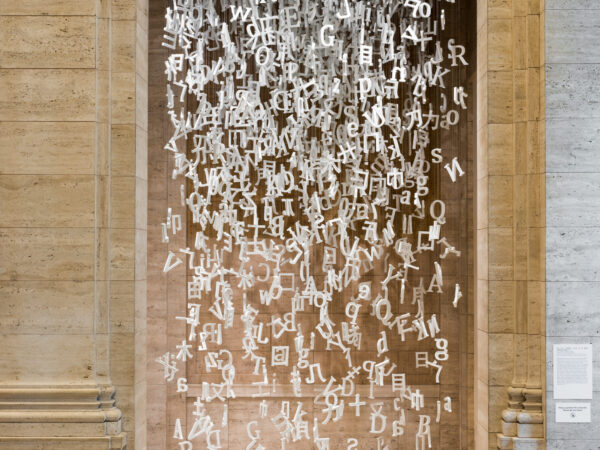 Image of white porcelain letters and parts of Chinese characters hanging from strings from the ceiling seen through a stone archway.