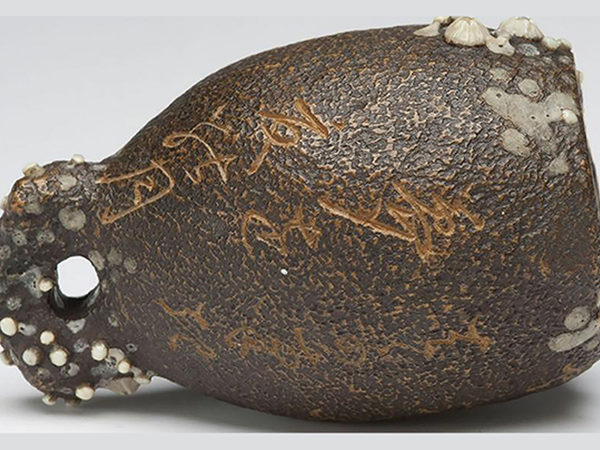 Thumb-sized carving of a brown, bell-shaped vessel covered with white barnacles and inscribed with cursive Japanese calligraphy in gold.