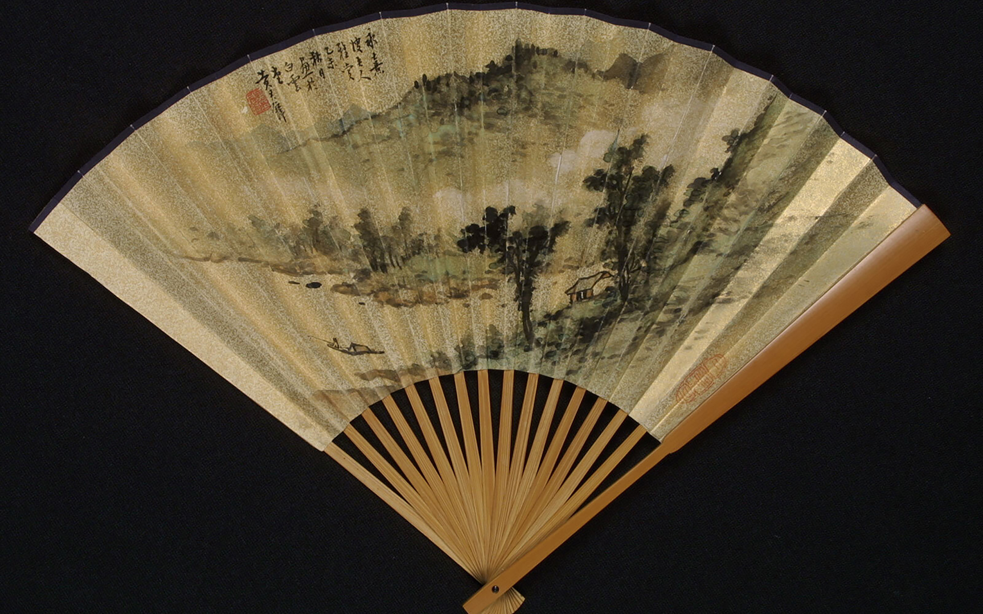 A folding paper fan spread open to reveal a landscape painted in black ink and washes of blue, green, and brown, featuring a fisherperson in a small boat in the water at lower left, trees and small hut at right, mountains and mist receding into the distance. Nine vertical lines of Chinese characters in black and an artist’s seal in red is in the top left; another red seal is stamped along the lower right edge.