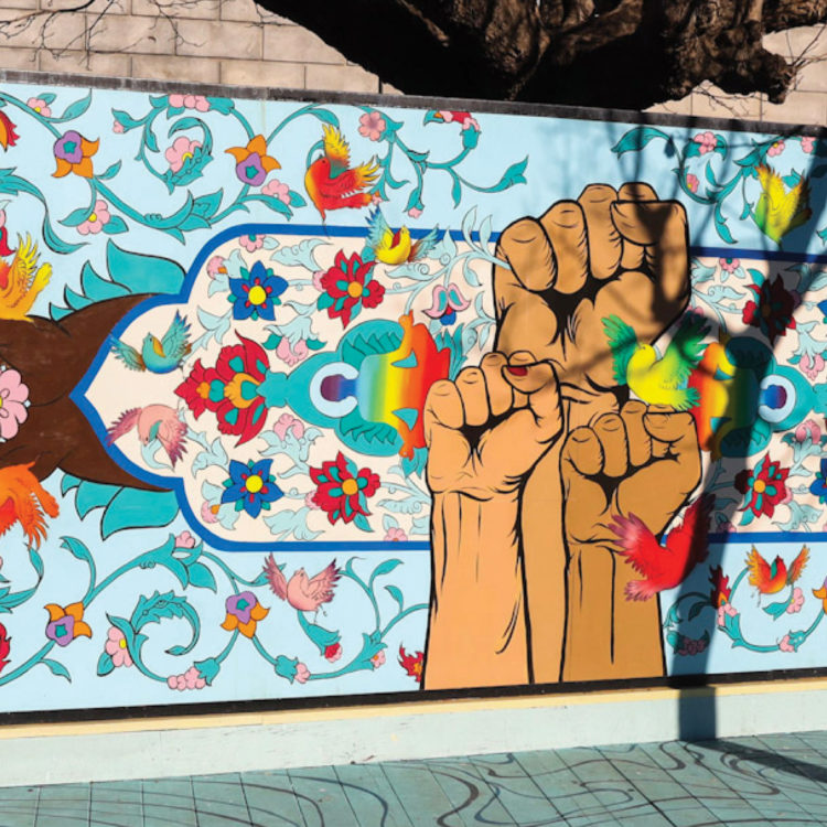 Image of a mural with a bright blue background, snake-like vines, colorful flowers and birds, and women with long, black hair dancing. In the center of the mural, three fists framed by a horizontal rectangular shape are raised in a power salute. In the rectangle are more colorful flowers and birds. Two hands cupping flowers stick out of both ends of the rectangle.