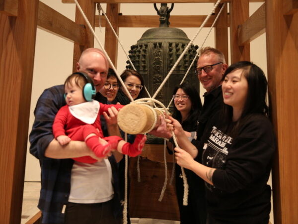 A group of six adults standing in front of a larger, Japanese temple bell. All are smiling, one is holding a baby in his arms.