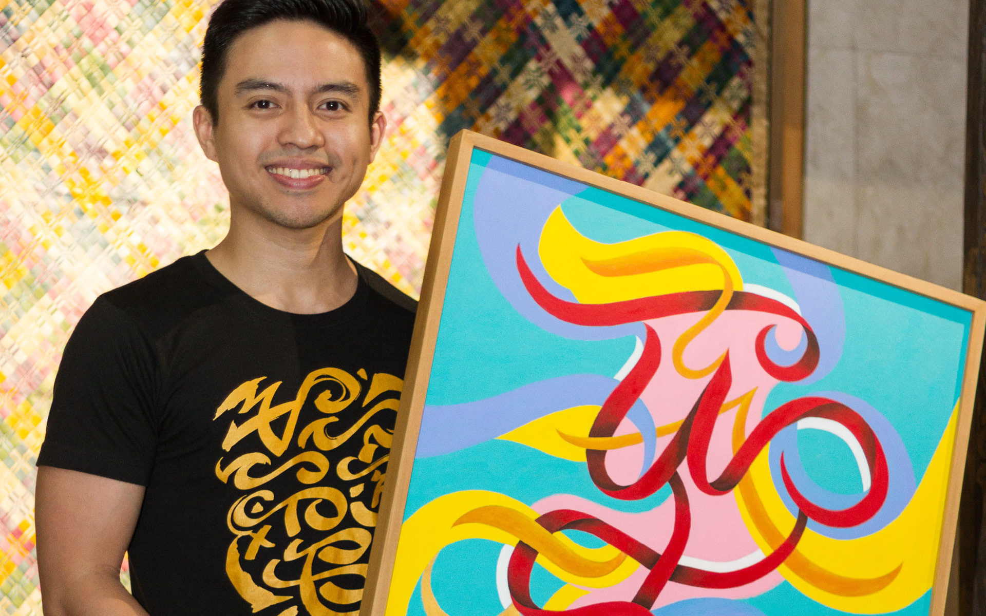 A smiling young man holds a 2 x 4' painting of a stylized script. The background is turquoise blue and the script is in pink, yello, red, and purple swirls.