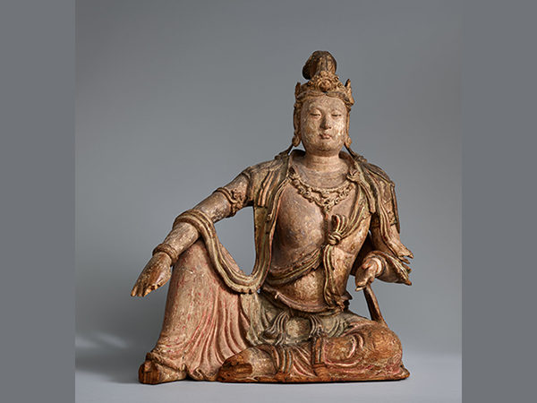 Larger than lifesize wooden crowned and bejeweled figure seated with right foot on the ground and right arm resting across the knee.