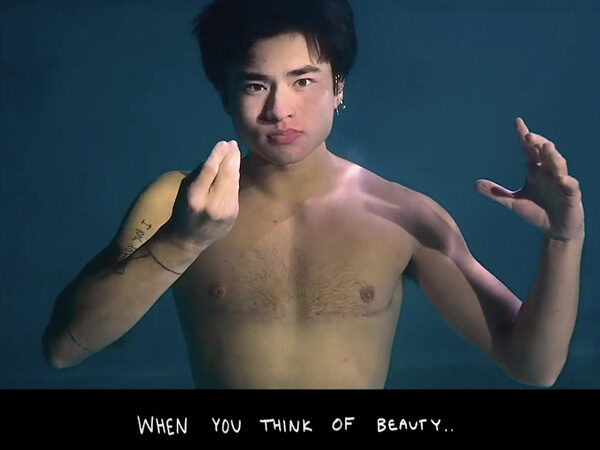 Chellaman, who is an Asian American Deaf transman poses underwater, his black hair floating above his head, and his hand making an ASL sign in front of his chest, where is top surgery scars can be seen. Text reads: “When you think of beauty…”