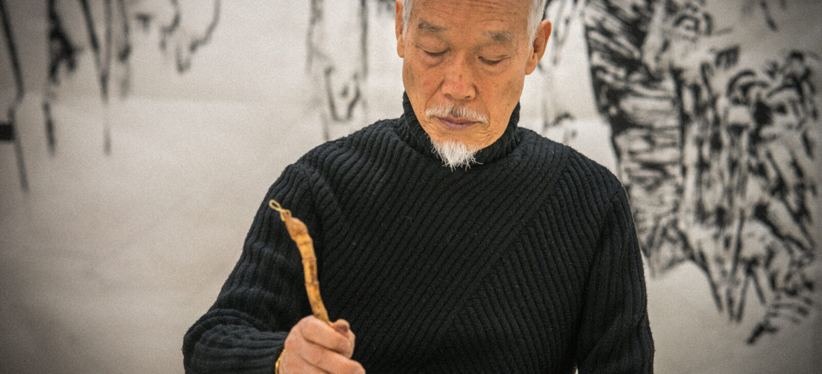 Artist Park Dae Sung holds an ink brush with a natural curving handle. He wears a black turtleneck sweater. Behind him is a soft focus ink painting of nature.