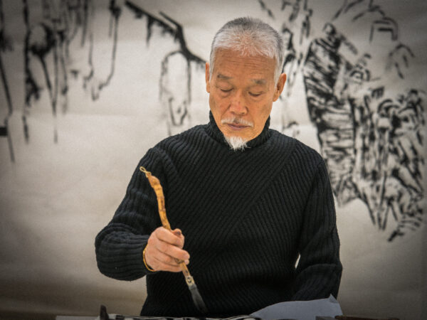 Artist Park Dae Sung holds an ink brush with a natural curving handle. He wears a black turtleneck sweater. Behind him is a soft focus ink painting of nature.