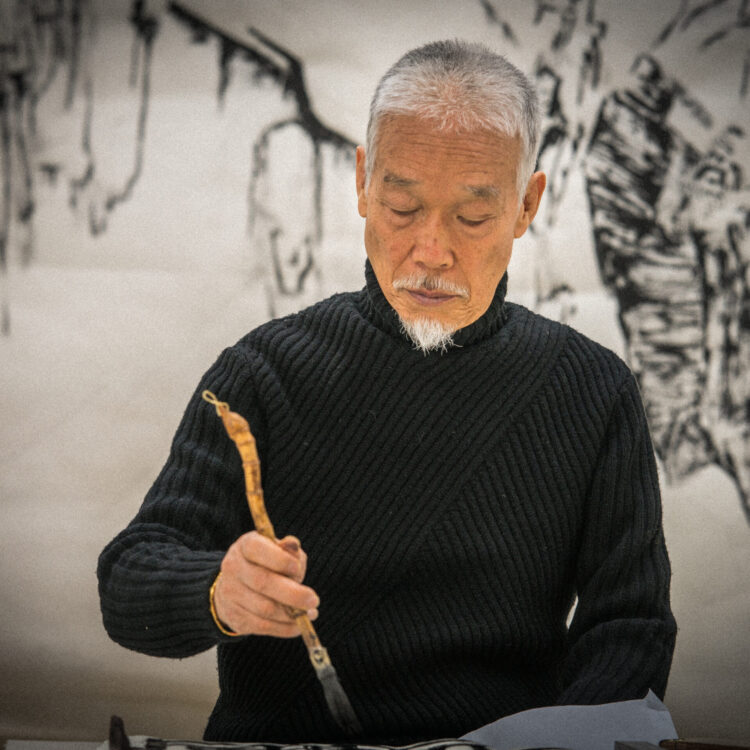 Artist Park Dae Sung holds an ink brush with a natural curving handle. He wears a black turtleneck sweater. Behind him is a soft focus ink painting of nature.